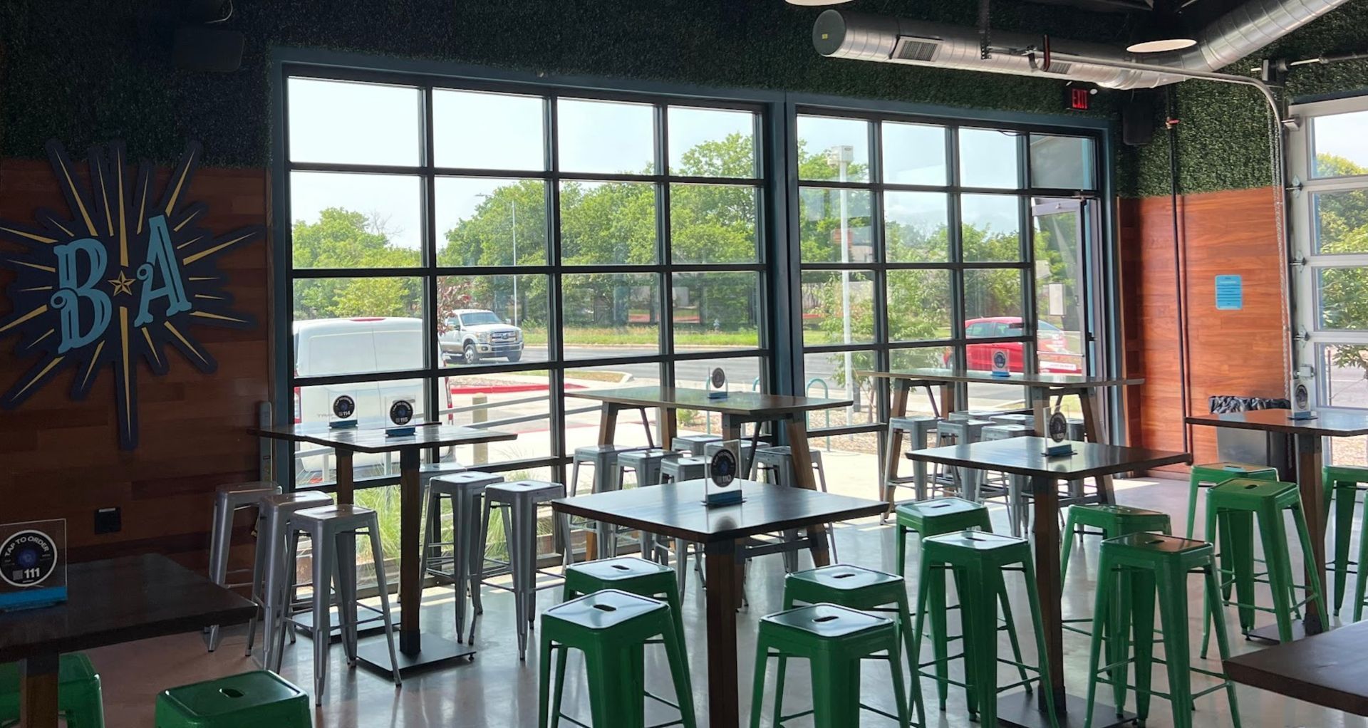 An indoor bar area with multiple tall tables and green stools, featuring large glass garage-style doors opening outside.
