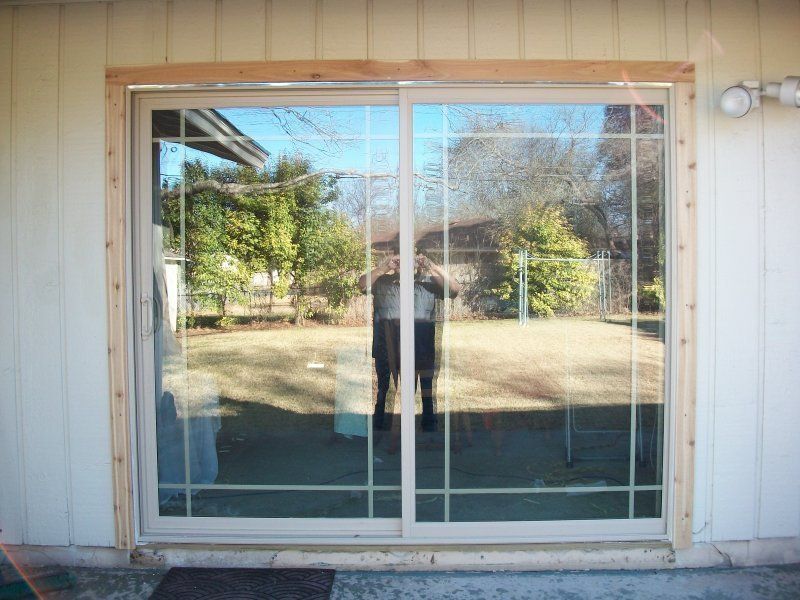 A sliding glass door with a beige frame installed in a light-colored wood-paneled wall, reflecting the yard outside.