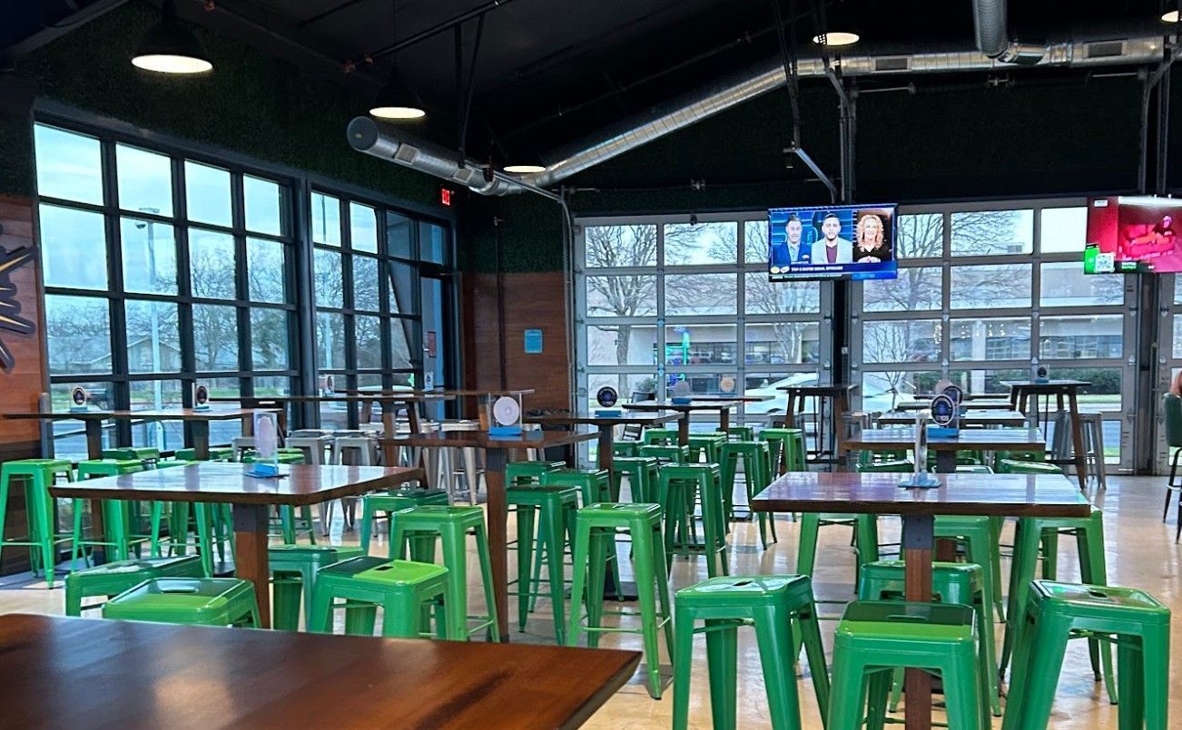 An indoor bar area with multiple tall, wooden tables and bright green metal stools under high ceilings and large windows.