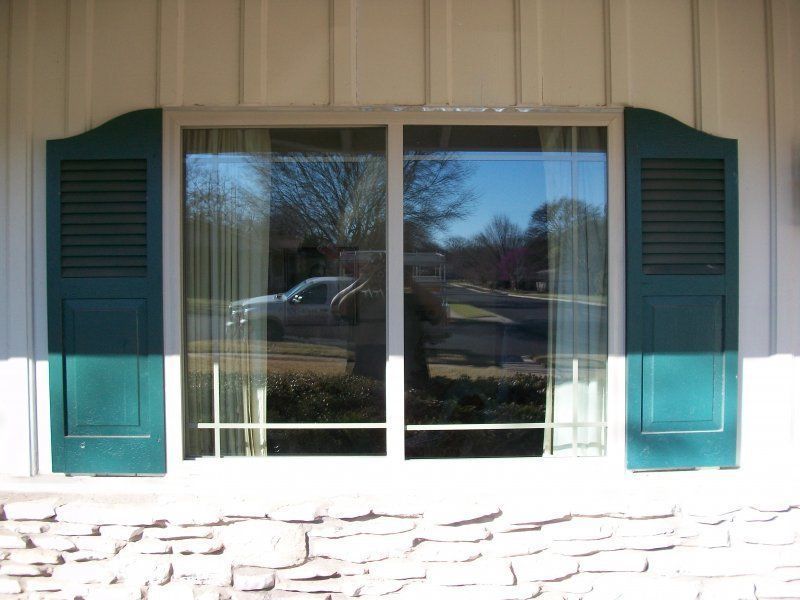 A rectangular window with dark green shutters on a house wall with stone-textured siding.