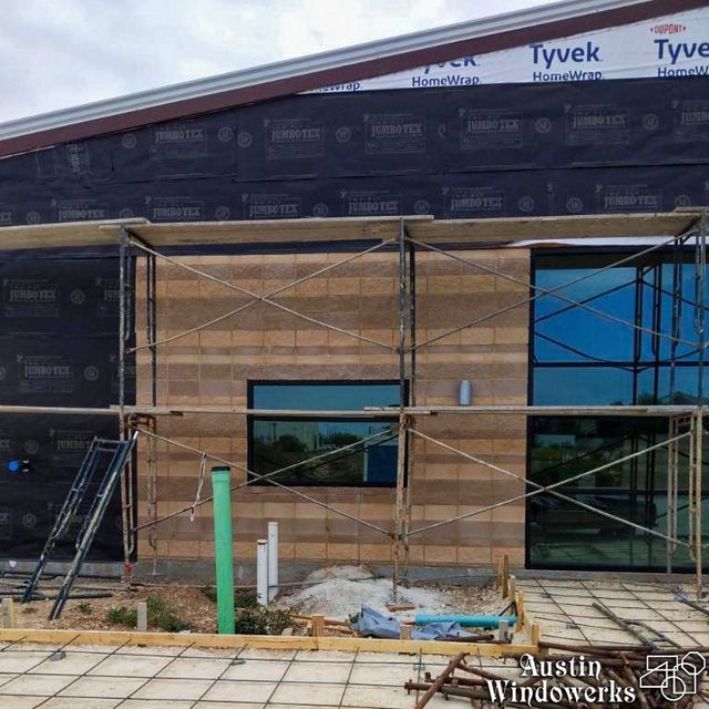 Scaffolding stands before a building under construction featuring beige masonry, dark glass windows, and Tyvek house wrap.