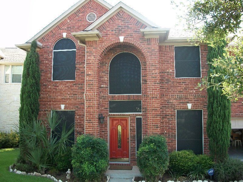 A two-story red brick house with arched windows covered in black sun screens and a prominent red front door.