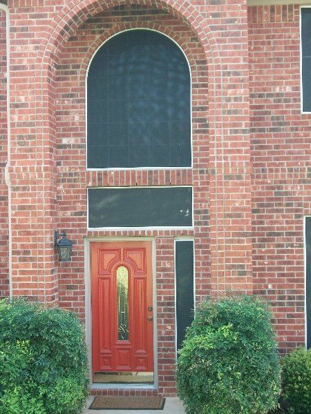 A red front door centered in a brick home entryway with a large arched window above it and two bushes in front.