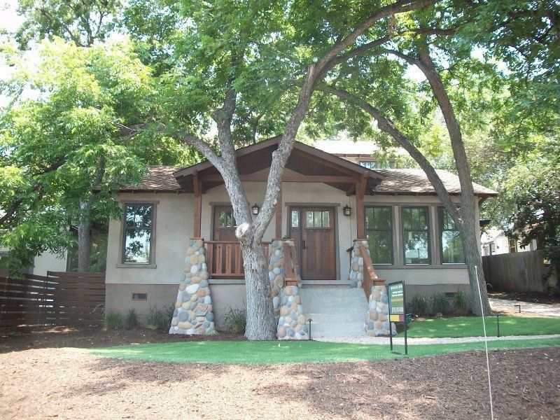 A tan house with stone porch columns, a wood front door, and large mature trees in the front yard.