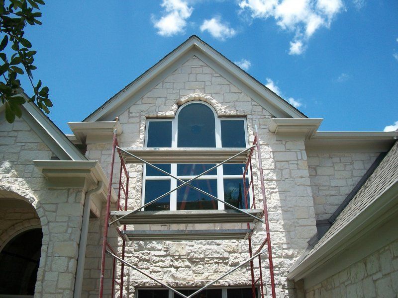 Scaffolding stands in front of a stone house facade with a large arched window under a gabled roof against a blue sky.