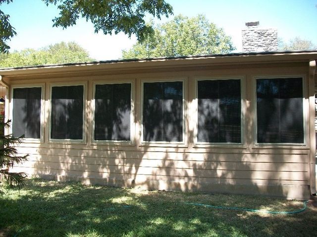 A tan exterior wall of a house featuring six rectangular, dark-screened windows, situated outdoors near trees and grass.