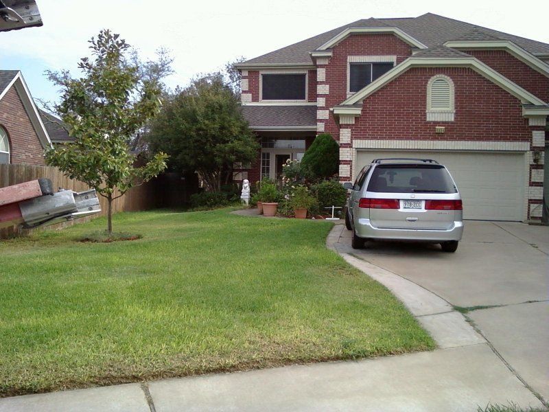 A silver minivan parked in the driveway of a two-story red brick suburban house with a front lawn and trees.