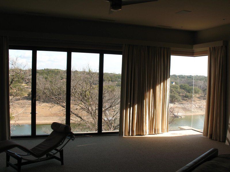 A room with a large wall of windows overlooking a lake, featuring a lounge chair and beige curtains.