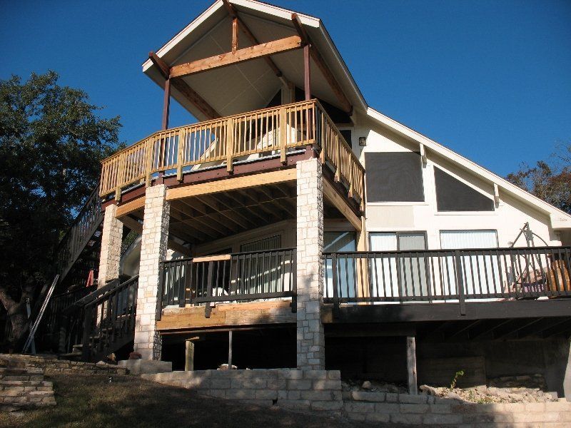 A two-story house with a wooden upper deck and a stone-pillared lower deck, situated on a grassy slope under a clear sky.