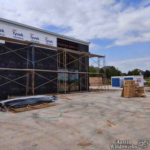A building under construction with scaffolding against a dark exterior wall and Tyvek wrap, with materials on the ground.