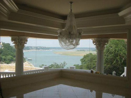 A room with a glass-enclosed balcony and ornate columns overlooking a wide river with trees and a sandy bank.