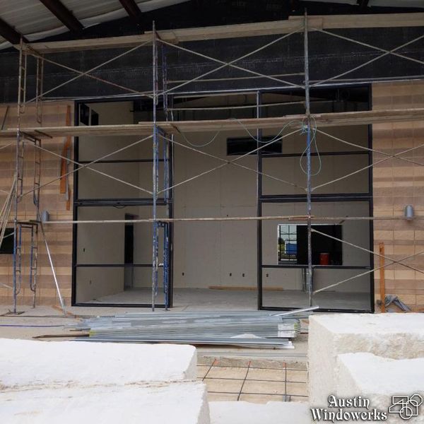 Construction site showing a large, rectangular black-framed glass storefront installation under scaffolding.