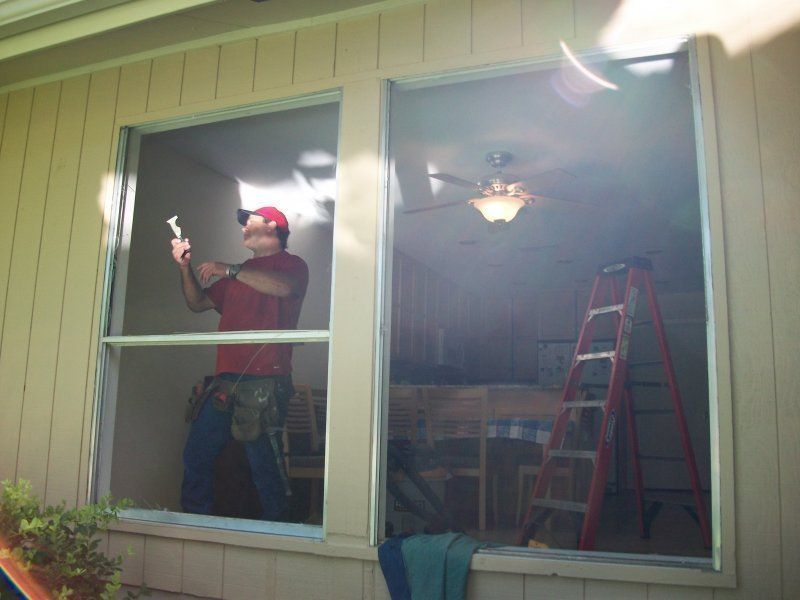 A person in a red shirt and tool belt works on a window frame, with a red ladder inside the room behind them.
