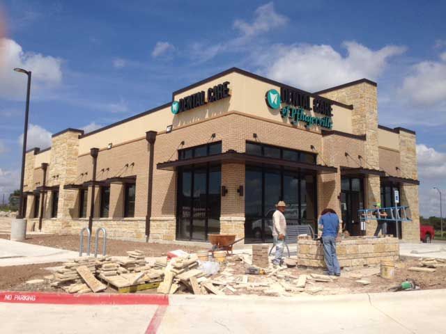 Two construction workers assemble a stone wall in front of a newly built Urgent Care facility under a sunny blue sky.