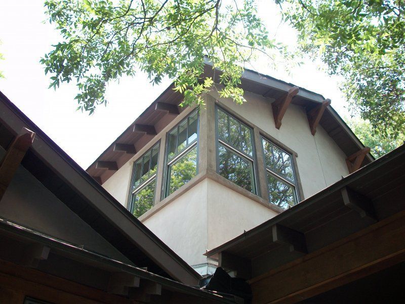 A low-angle view of a multi-story house with light-colored stucco walls, wooden trim, and windows framed by trees.