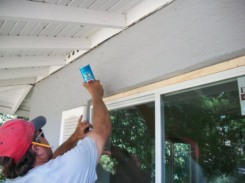 A worker in a red cap applies expanding foam sealant to the gap above a newly installed window on an exterior wall.
