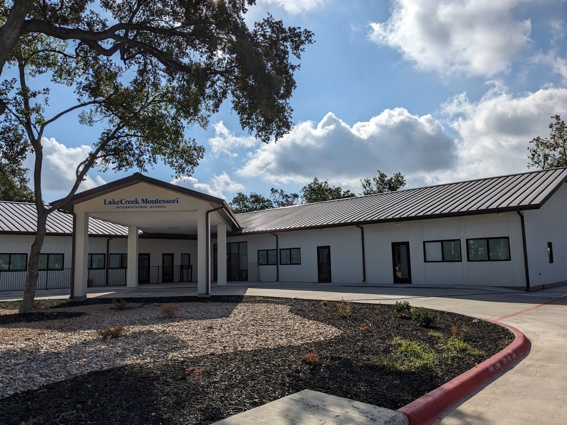 A single-story white building with a covered entrance and metal roof surrounded by landscaping under a blue, cloudy sky.