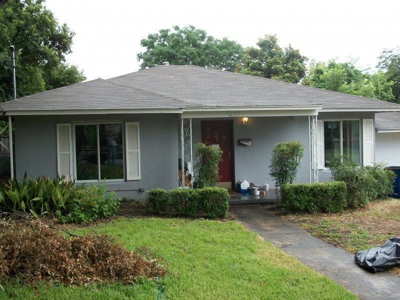 Single-story gray house with a dark roof, white shutters, front porch, and trimmed hedges in a grassy front yard.