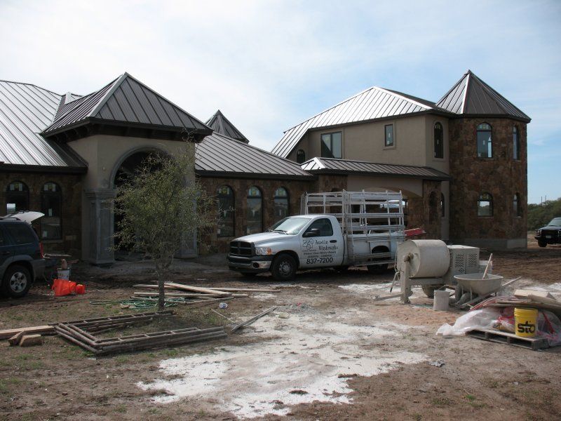 A multi-story stone house with a metal roof under construction, featuring a work truck and concrete mixer in the yard.