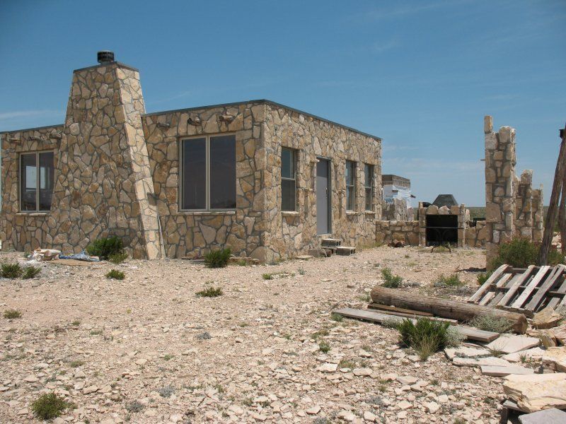 A stone house with a prominent chimney stands in a dry, rocky desert landscape under a clear blue sky.