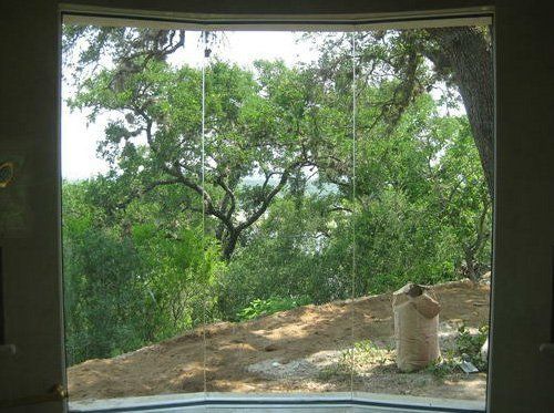 A large window frames a view of lush green trees and a sloped, dirt-covered yard outside.