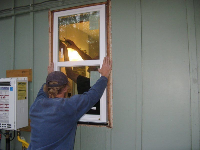 A person in a blue long-sleeved shirt installs a white-framed window into the green exterior wall of a building.
