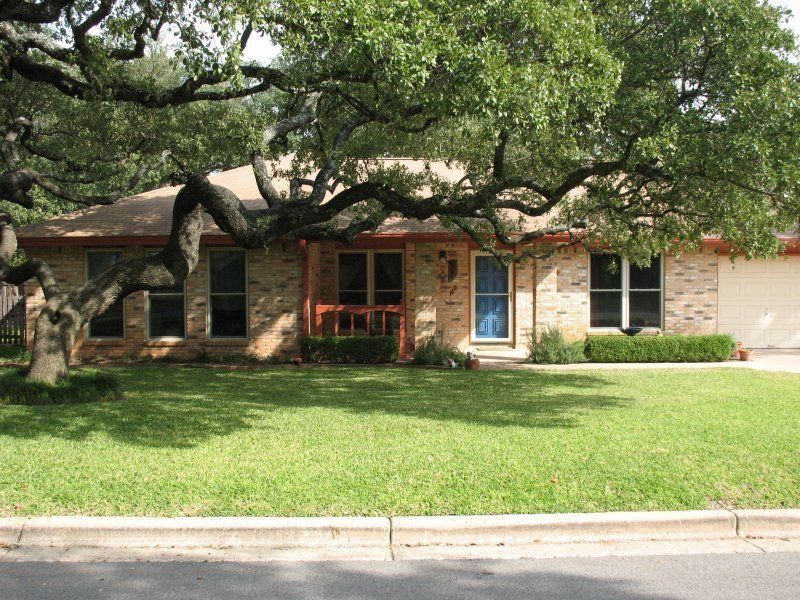 A single-story brick house with a blue front door, tan roof, and a large oak tree spreading across the front lawn.