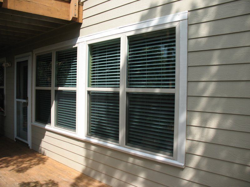 A large, multi-pane rectangular window with white trim on a beige-sided house next to a door on a wooden deck.