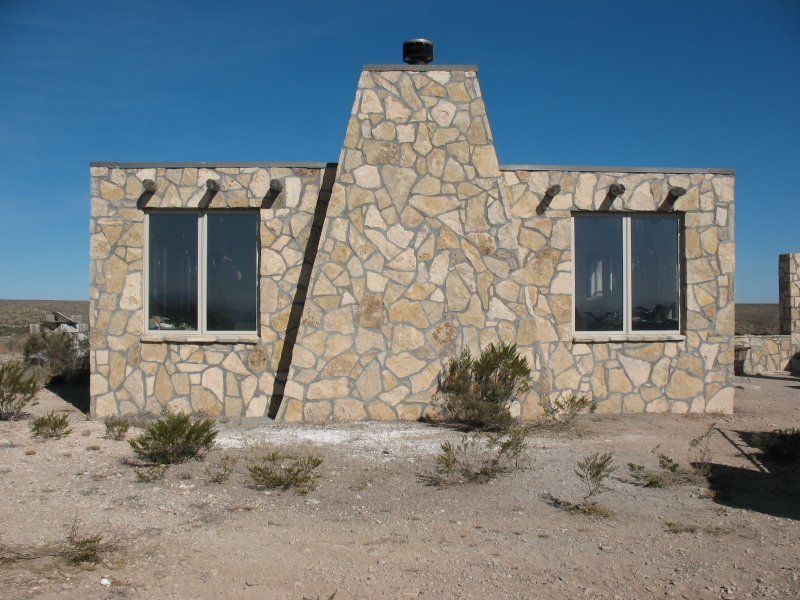 A stone house with two windows and a central chimney set in a desert landscape under a clear blue sky.