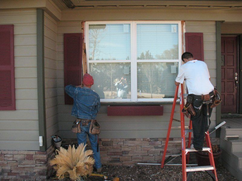 Two workers in work gear install a window in the exterior wall of a house with stone siding.