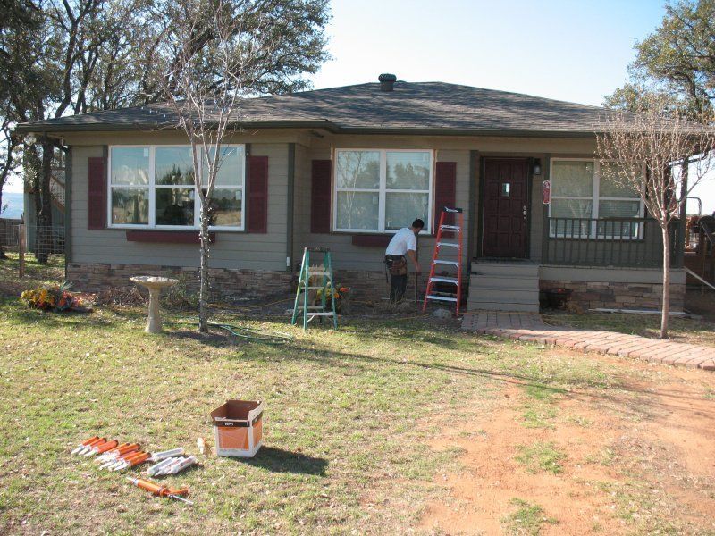 A worker uses a ladder to paint the exterior of a tan house with maroon shutters, surrounded by trees and tools.