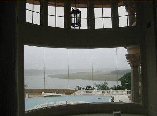 A view through a large curved window overlooking a swimming pool, a stone balustrade, and a lake under a cloudy sky.