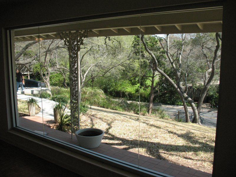 A view through a large window showing a grassy hillside with trees, a stone patio, and a decorative white post.