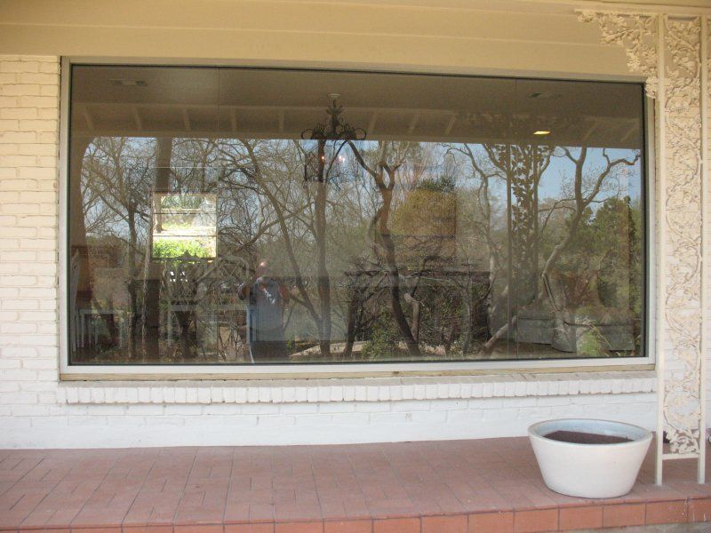 A large, rectangular window on a white brick wall, reflecting trees and a patio, with a white planter on the porch floor.
