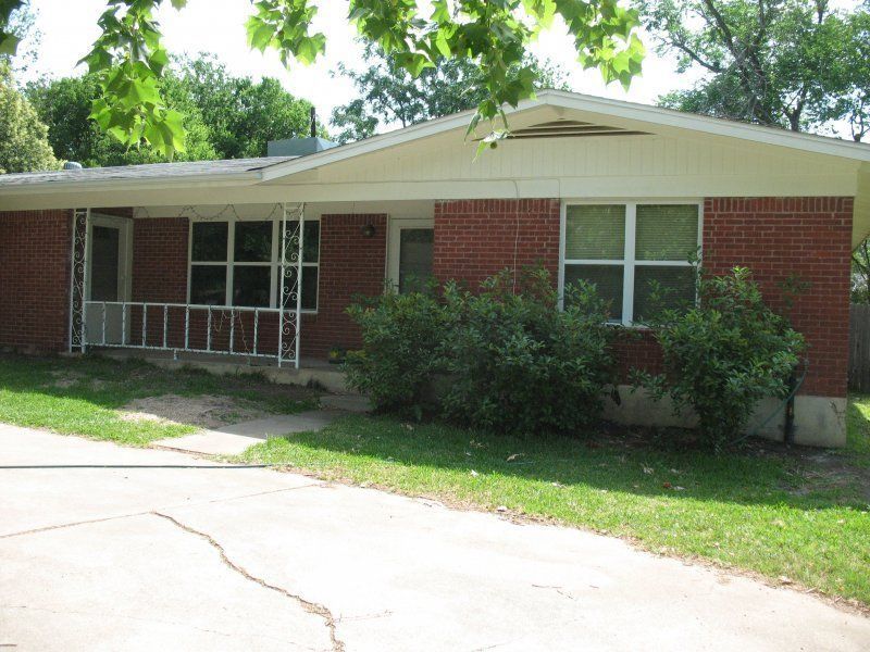 Single-story red brick house with white trim, a porch with metal railings, and a concrete driveway under leafy trees.