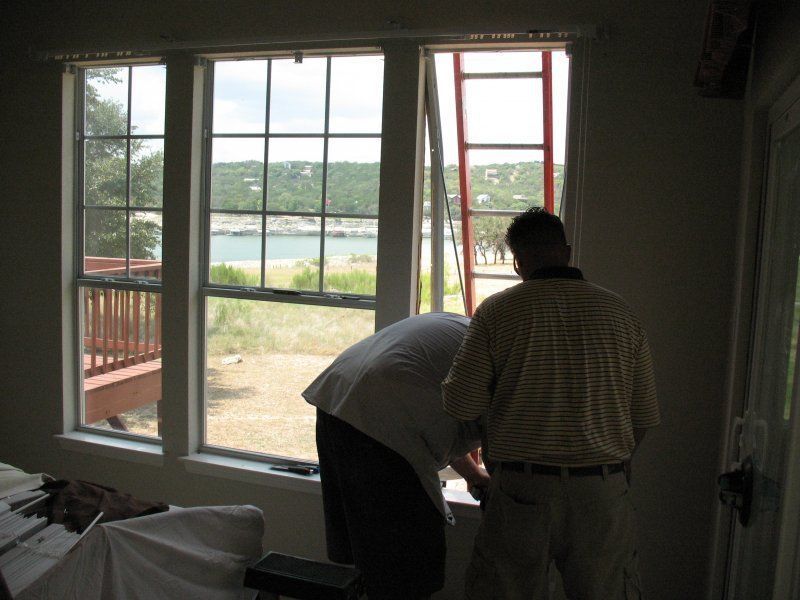 Two people work on a window frame in a room, with a ladder leaning against the glass and a lake view in the background.