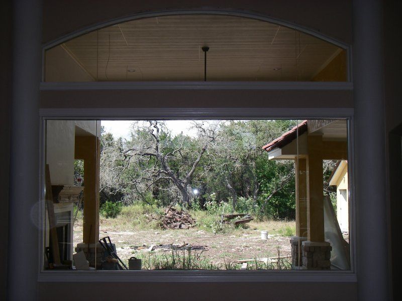 View through a large rectangular window and an arched transom looking out onto a wooded yard with an unfinished patio.