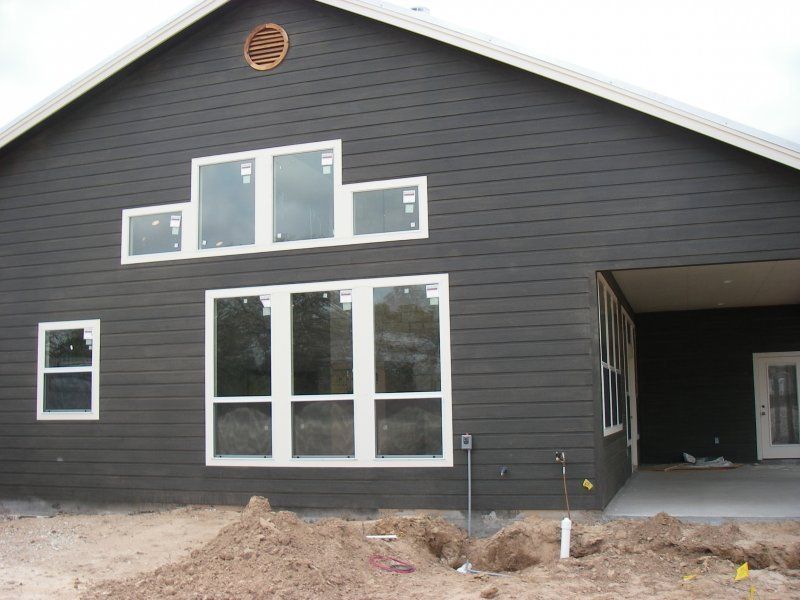 Dark grey house exterior with light trim, large window groupings, a gable vent, and a covered porch under construction.
