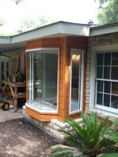 A bay window with cedar wood paneling is installed on the side of a light-colored stone house near a garden.
