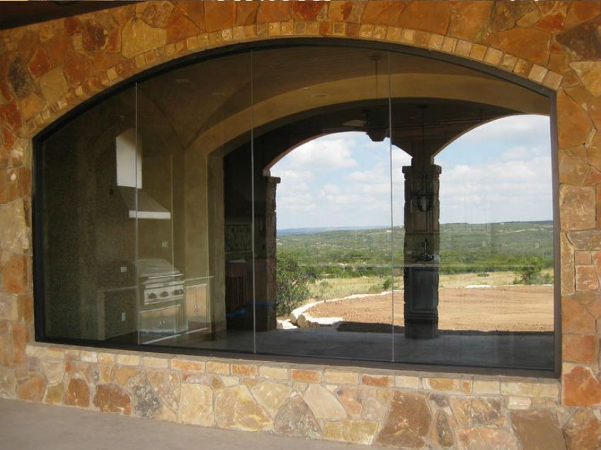 A wide, arched window with a black frame shows a reflection of a stone patio, an outdoor kitchen, and a landscape view.