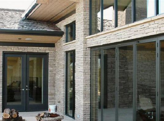 Modern stone exterior of a home featuring black-framed double doors and a glass wall, with a small outdoor fire pit.