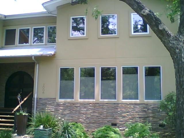 A two-story house with tan stucco, stone veneer siding, and multiple rectangular windows, partially shaded by a tree.