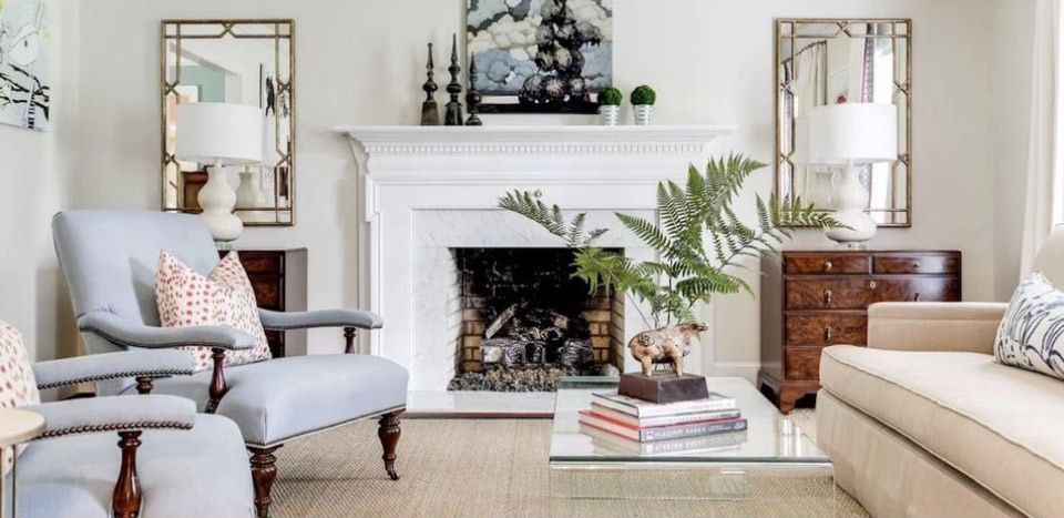Elegant living room with white fireplace, mirrored decor, beige sofa, armchair, and coffee table with books and greenery.