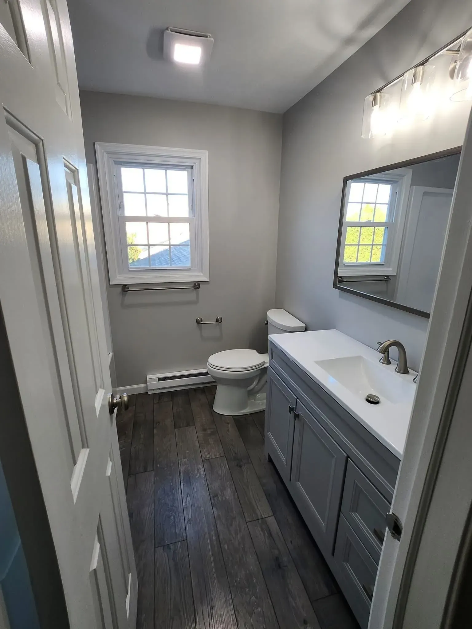 Narrow gray bathroom with white vanity, toilet, window, mirror, and dark wood floor.