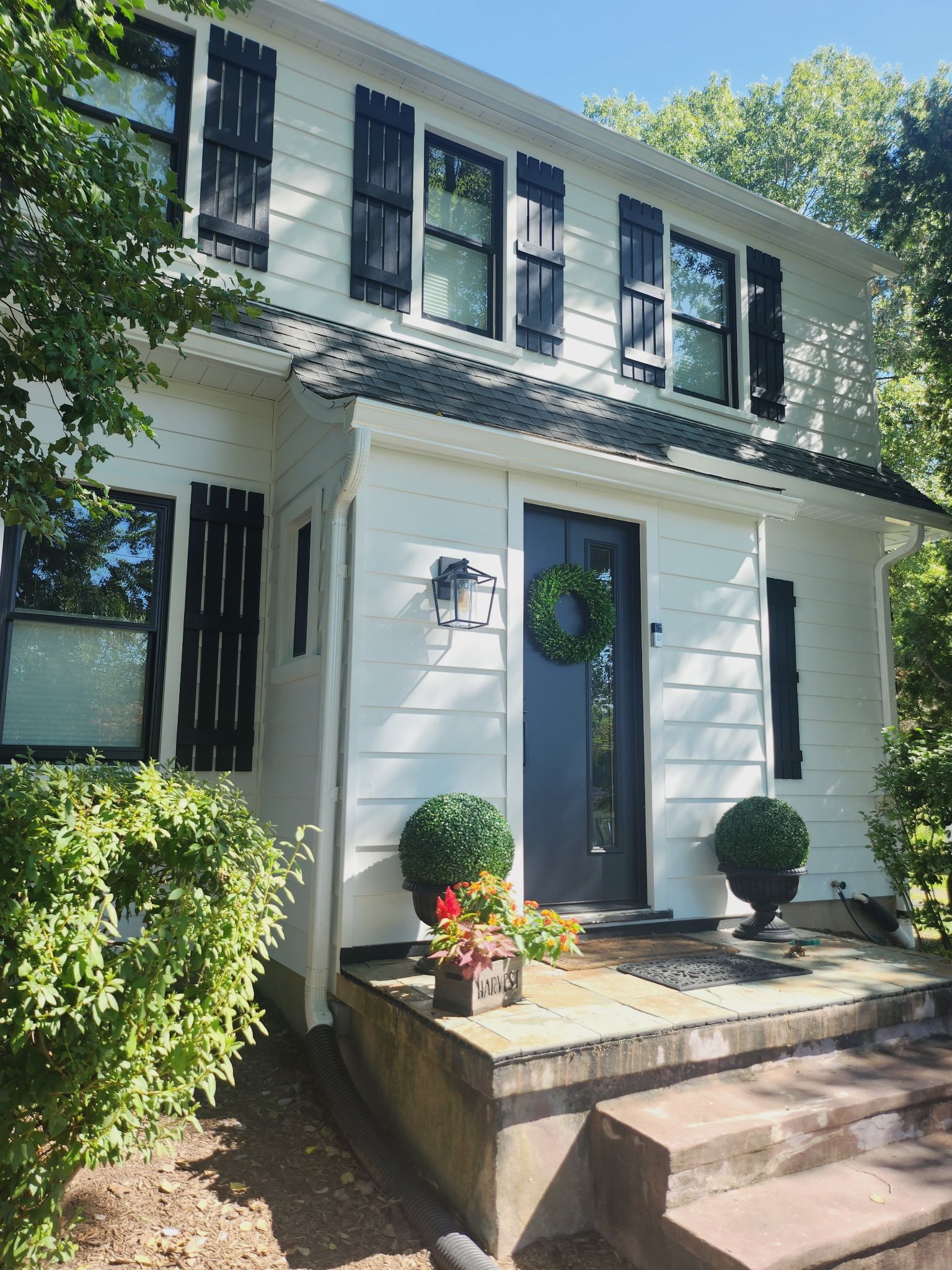 Front entrance of a white two-story house with blue shutters, a wreath, and potted plants on the steps