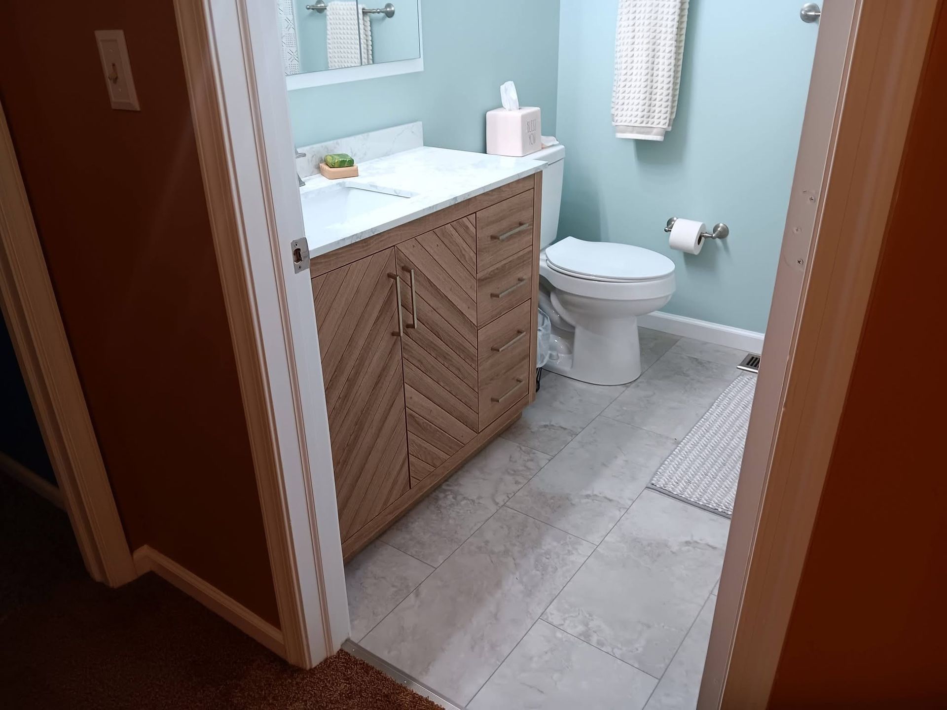 Small bathroom with wood vanity, white sink and toilet, light blue walls, and gray tile floor.