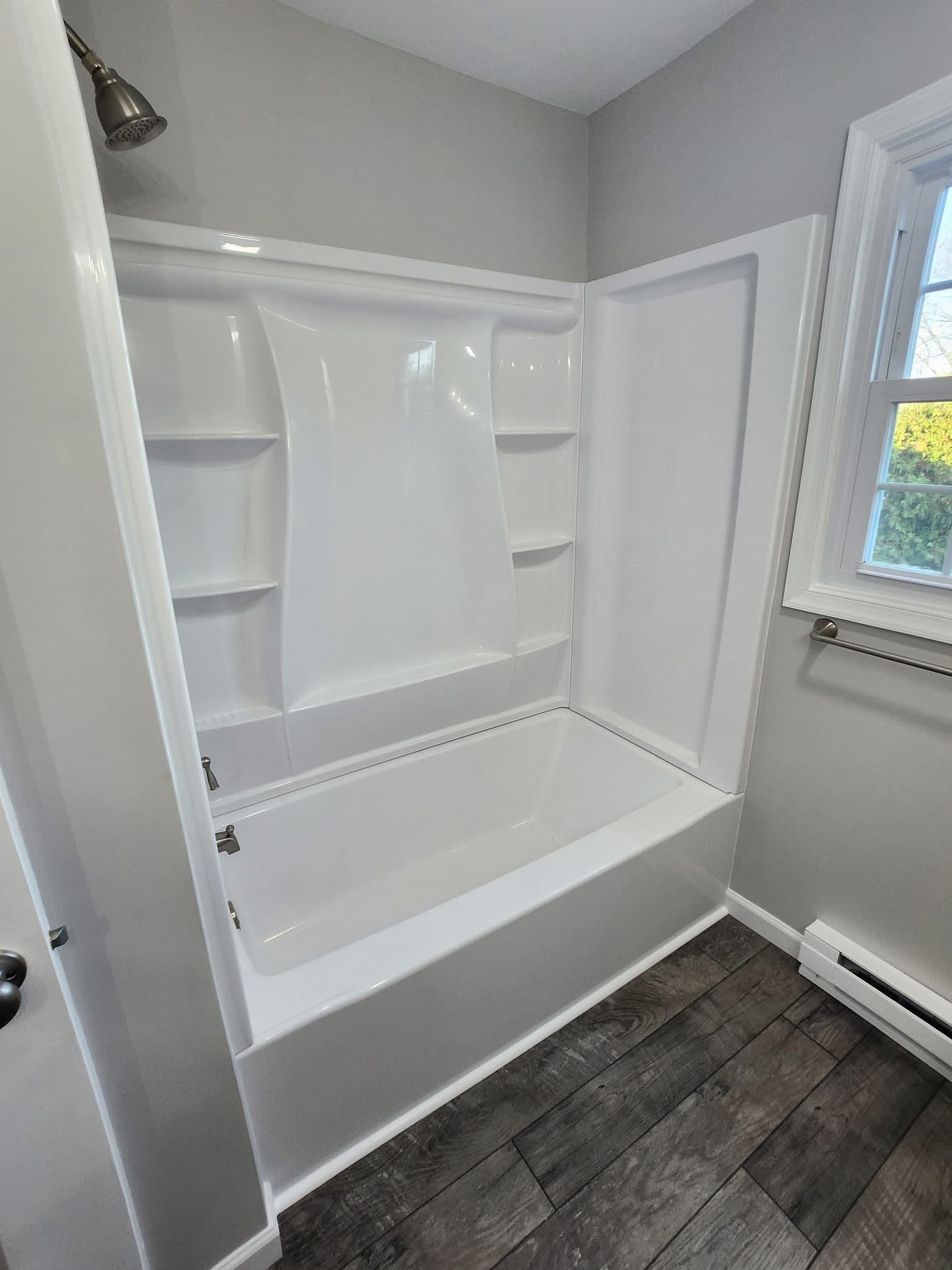 White bathtub with built-in shelves in a small bathroom with dark wood-style floor and window light
