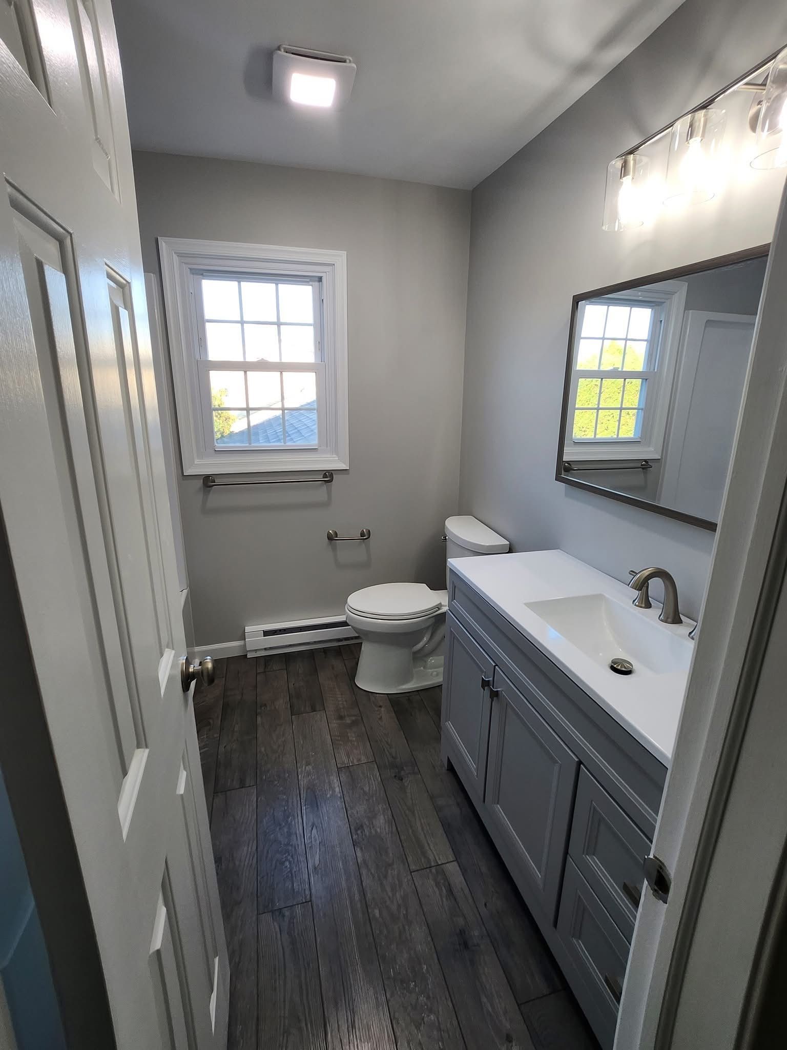 Small bathroom with gray vanity, white sink, toilet, window, and dark wood floor.