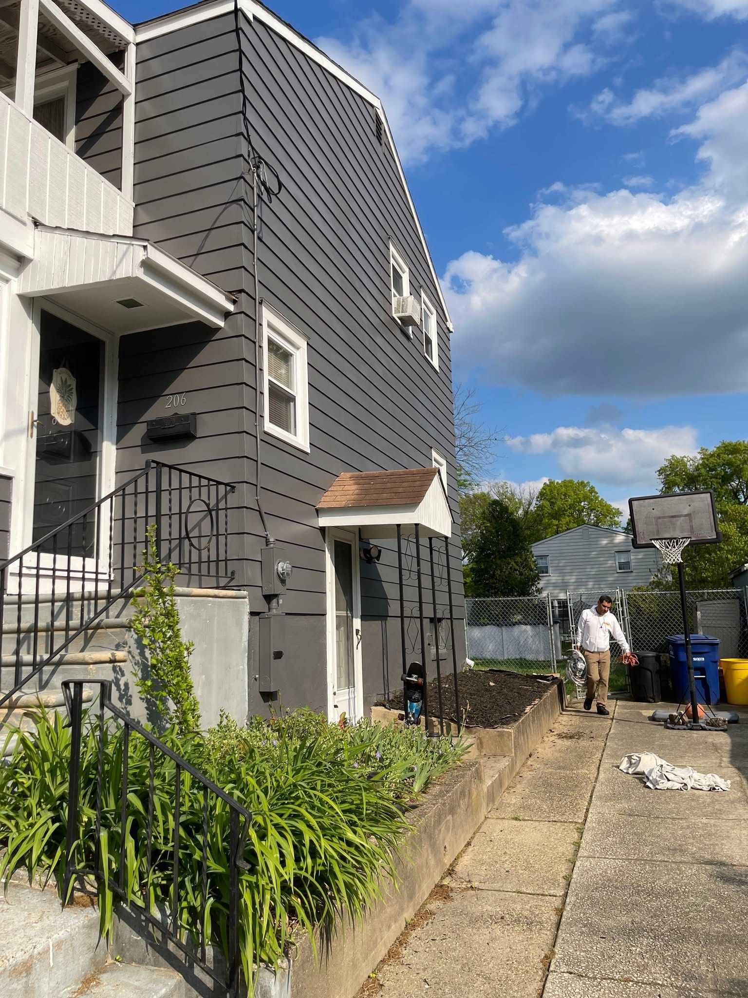 Narrow side yard beside a gray house with stairs, railing, plants, and a gravel path under a blue sky.