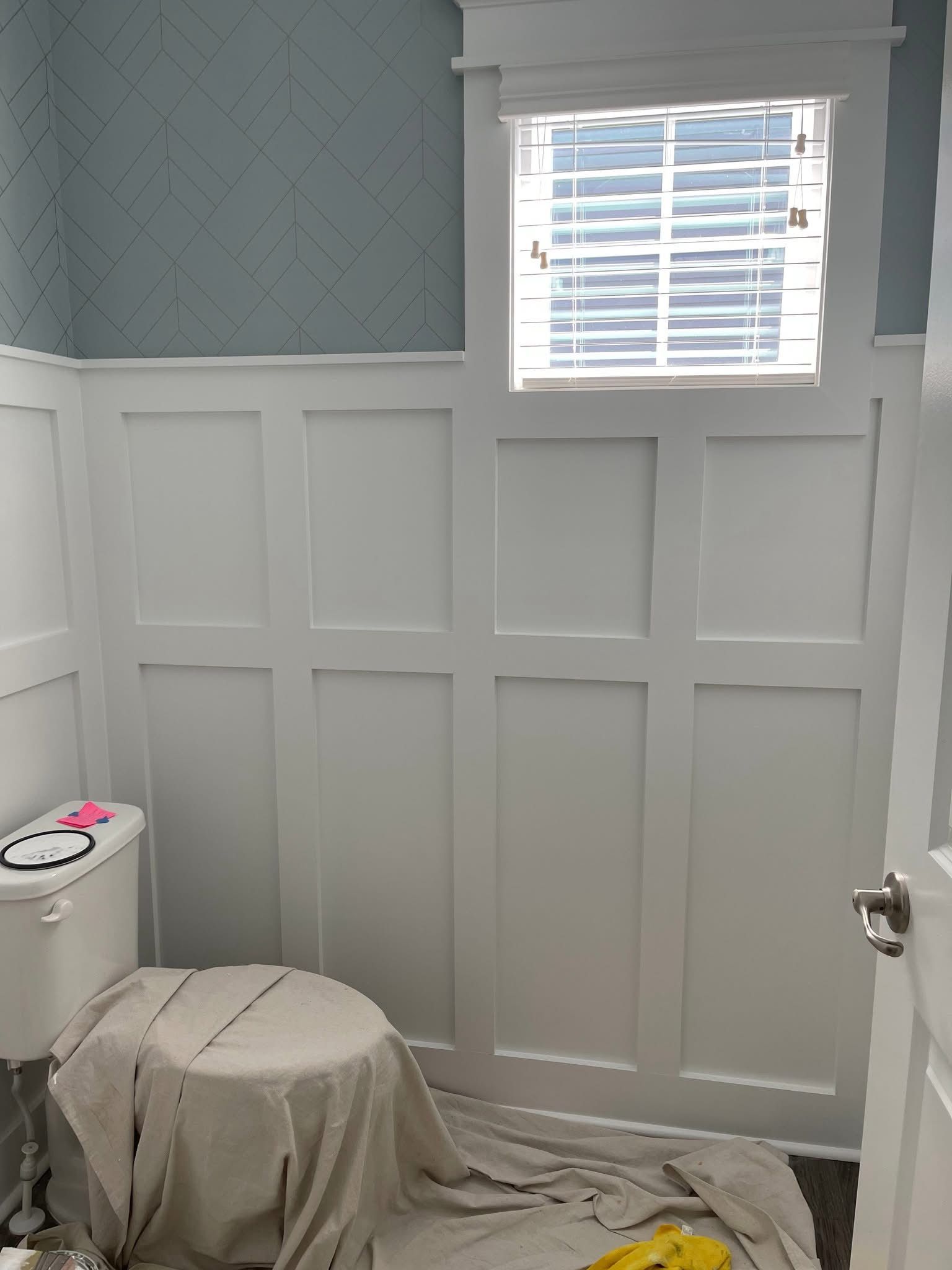 Small bathroom with white wainscoting, blue patterned wallpaper, and a window above a toilet.